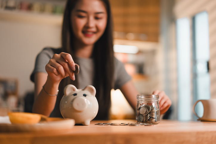 Saving money is important habit, as shown by young woman joyfully depositing coins into piggy bank while surrounded by jar of coins. This scene captures essence of financial responsibility and